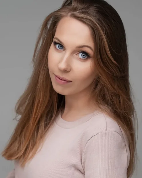 Eliza Klucz – portret: długie, brązowe włosy, jasnoróżowy sweter, szare tło
